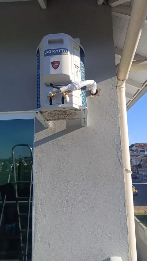 Condensadora externa Agratto Inverter branca fixada em parede texturizada cinza, com tubulação de cobre isolada entrando na alvenaria