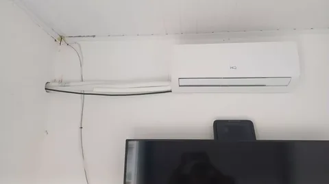 Evaporadora split HQ branca instalada acima de televisão na sala, com tubulação frigorígena e cabo elétrico saindo em rasgo na parede