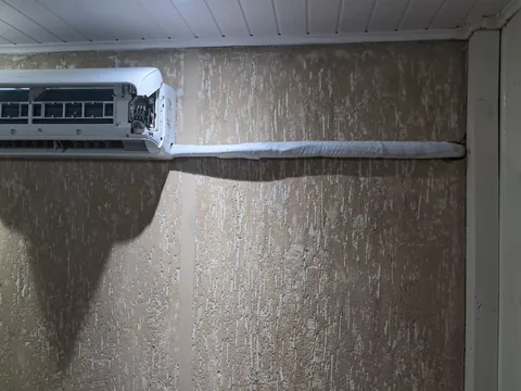 Unidade interna com painel lateral aberto e tubulação isolada em parede com textura bege (grafiato), sob forro branco